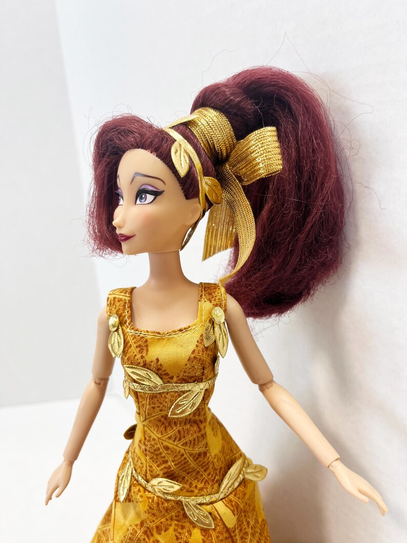 May include: A doll with dark reddish-purple hair styled in a high ponytail with a gold ribbon. The doll wears a yellow dress featuring a brown leaf pattern, accented with gold leaf-shaped embellishments on the bodice and waistline.
