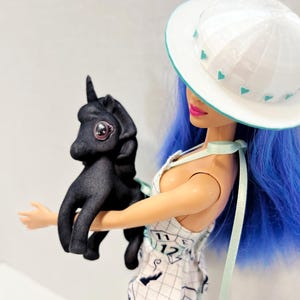 May include: A doll with bright blue hair wearing a white hat with teal heart accents and a white dress featuring a grid pattern with numbers and shapes. The doll is holding a small, matte black unicorn figurine with large, dark, glossy eyes.
