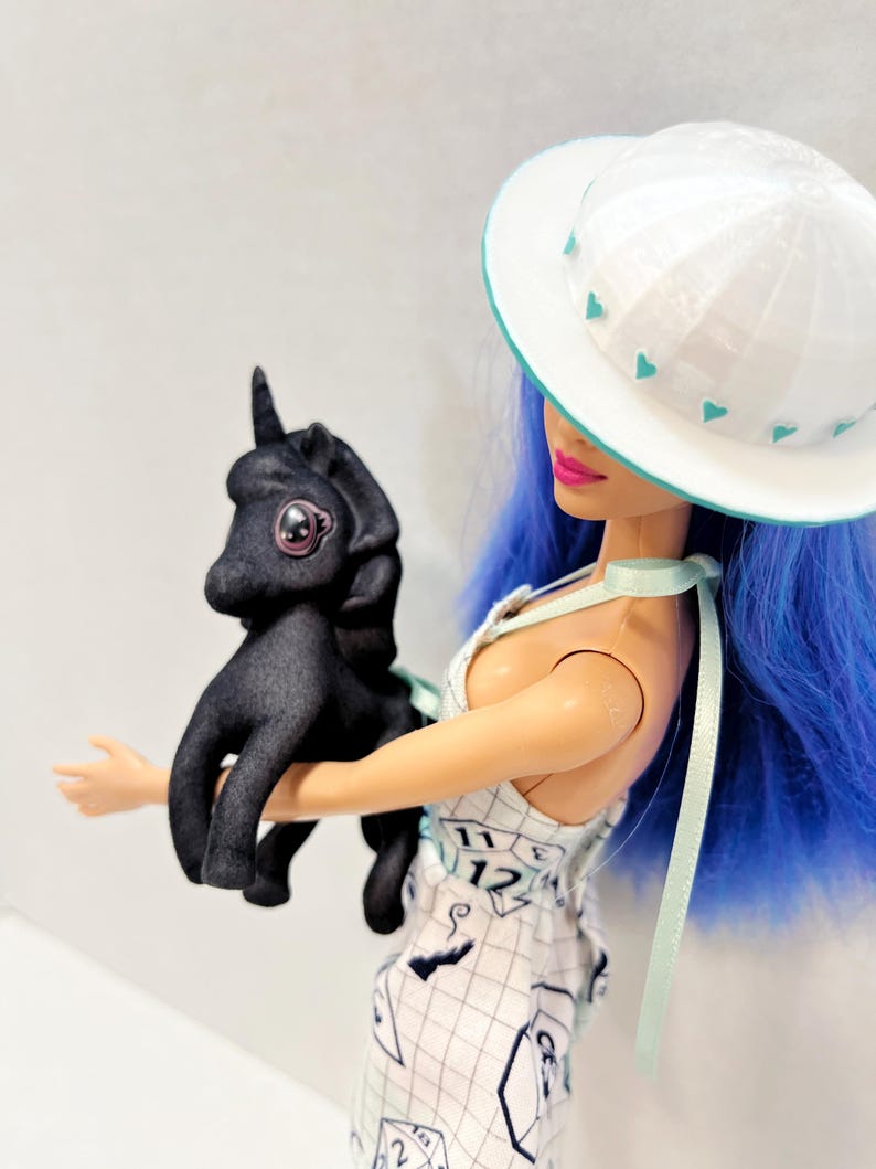 May include: A doll with bright blue hair wearing a white hat with teal heart accents and a white dress featuring a grid pattern with numbers and shapes. The doll is holding a small, matte black unicorn figurine with large, dark, glossy eyes.
