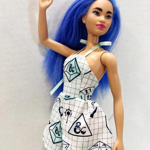 May include: A doll with long, vibrant blue hair and a light skin tone, wearing a white halter-style dress featuring a black grid pattern and various black and light green polyhedral dice graphics, including a twenty-sided die with the number 20 visible.