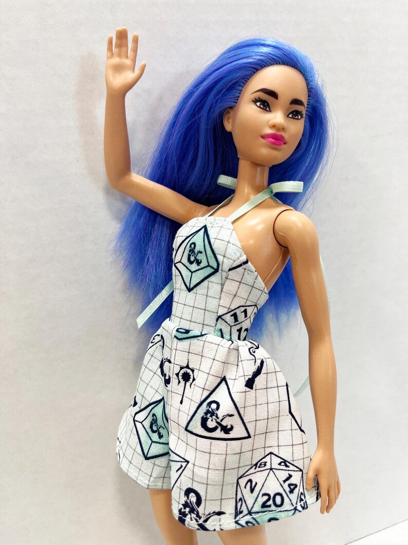 May include: A doll with long, vibrant blue hair and a light skin tone, wearing a white halter-style dress featuring a black grid pattern and various black and light green polyhedral dice graphics, including a twenty-sided die with the number 20 visible.