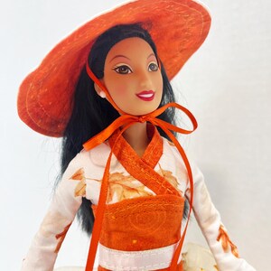 May include: A doll with long black hair wearing a wide-brimmed orange hat tied with a ribbon under the chin. The doll wears a white long-sleeved top featuring an orange leaf pattern, paired with an orange bodice and a layered skirt with tulle.