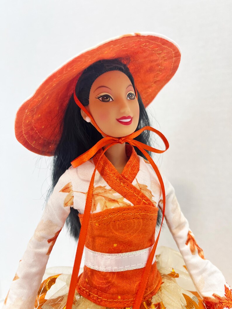 May include: A doll with long black hair wearing a wide-brimmed orange hat tied with a ribbon under the chin. The doll wears a white long-sleeved top featuring an orange leaf pattern, paired with an orange bodice and a layered skirt with tulle.