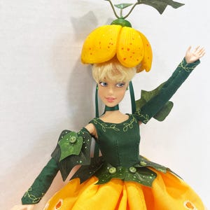 May include: A doll with blonde hair wearing a yellow flower-shaped hat with green leaves, a dark green bodice with embroidered vines, and a long yellow skirt featuring white eyelet embroidery. The doll has one arm raised and green leaf accents.