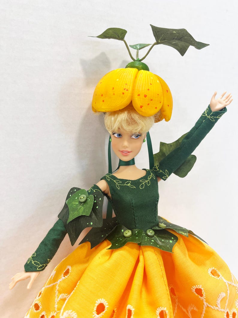 May include: A doll with blonde hair wearing a yellow flower-shaped hat with green leaves, a dark green bodice with embroidered vines, and a long yellow skirt featuring white eyelet embroidery. The doll has one arm raised and green leaf accents.