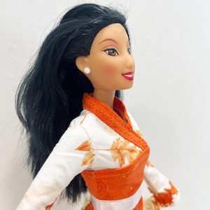 May include: A doll with long black hair, brown eyes, and red lips, wearing a white top featuring an orange leaf pattern, a solid orange wrap-style bodice, and a white ribbon waistband. The doll has small white stud earrings and rosy cheeks.