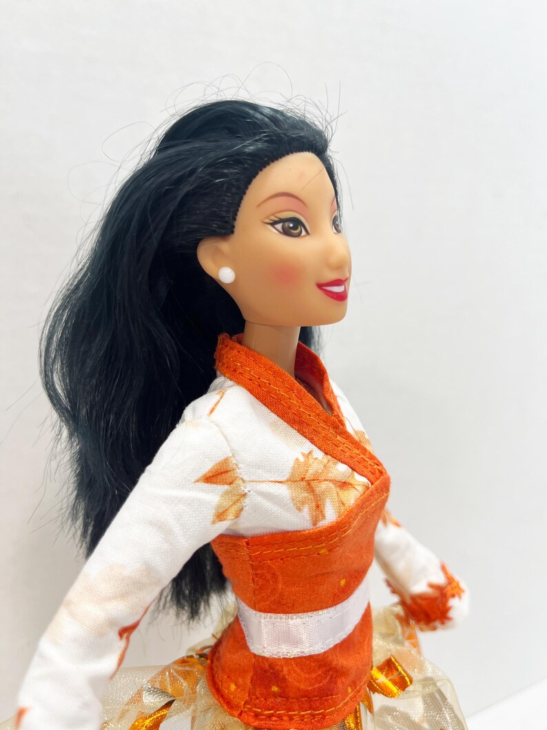 May include: A doll with long black hair, brown eyes, and red lips, wearing a white top featuring an orange leaf pattern, a solid orange wrap-style bodice, and a white ribbon waistband. The doll has small white stud earrings and rosy cheeks.