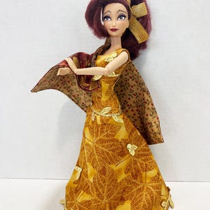 May include: A doll with dark red hair styled with a gold ribbon, wearing a yellow floor-length dress featuring a brown leaf pattern and gold leaf accents along the hem and waist. A matching brown and tan floral print shawl drapes over the shoulders.