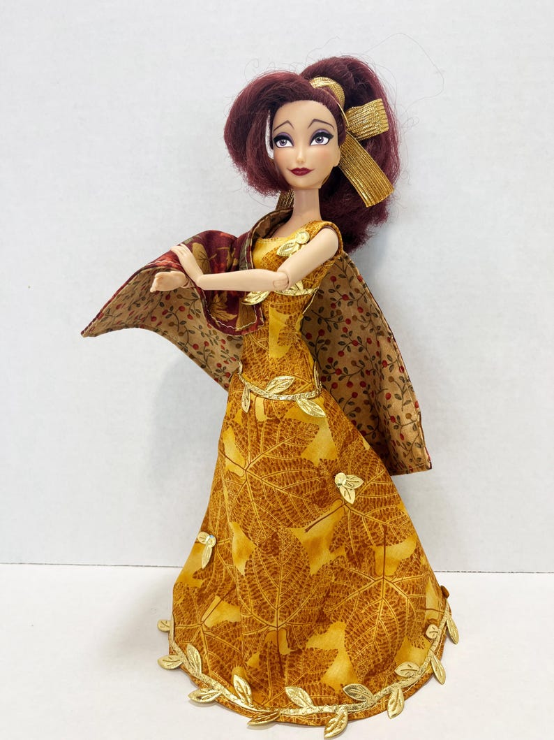 May include: A doll with dark red hair styled with a gold ribbon, wearing a yellow floor-length dress featuring a brown leaf pattern and gold leaf accents along the hem and waist. A matching brown and tan floral print shawl drapes over the shoulders.