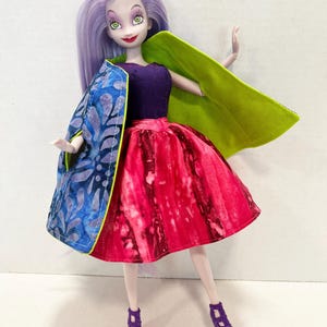 Pu&ograve; includere: Una bambola con lunghi capelli color lavanda, che indossa un corpetto viola scuro e una gonna tie-dye rosso e rosa vivace. La bambola indossa un mantello reversibile con un motivo a foglie blu e viola su un lato e verde lime sull'altro.