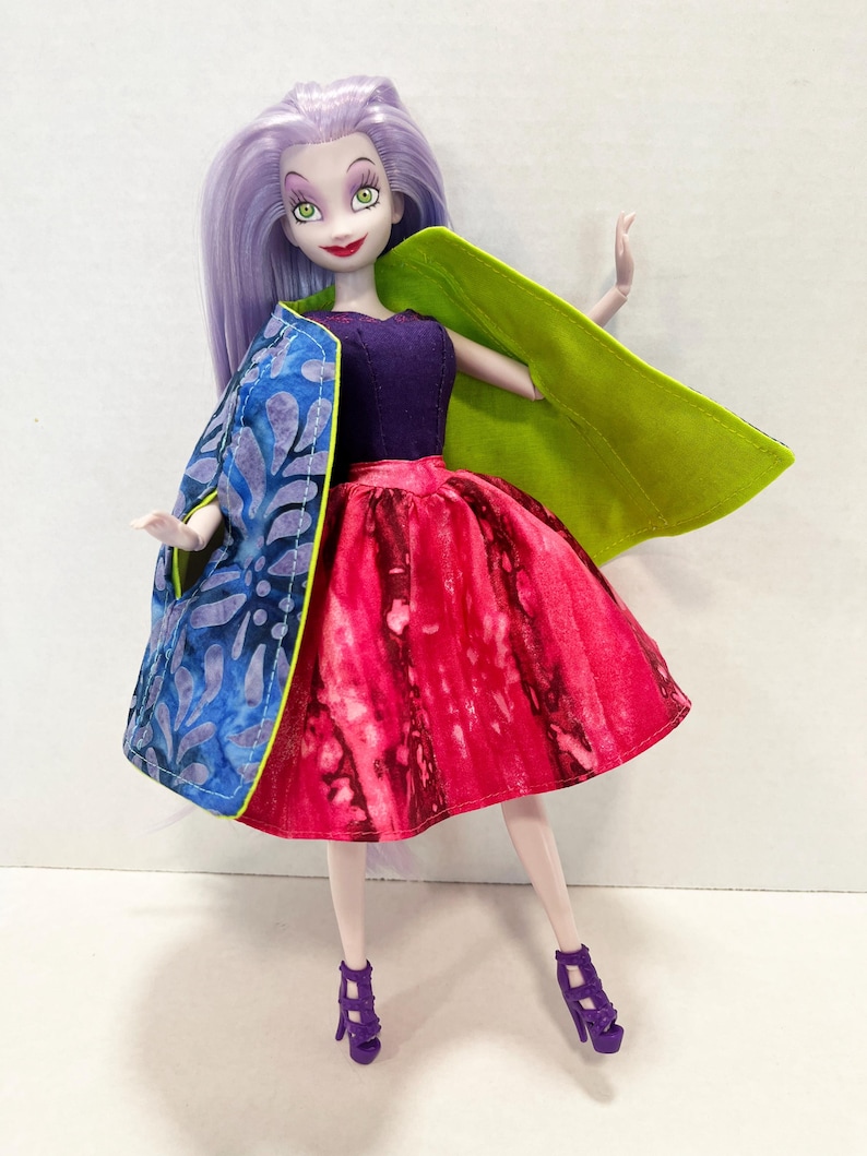 Pu&ograve; includere: Una bambola con lunghi capelli color lavanda, che indossa un corpetto viola scuro e una gonna tie-dye rosso e rosa vivace. La bambola indossa un mantello reversibile con un motivo a foglie blu e viola su un lato e verde lime sull'altro.