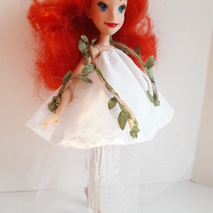 Puede incluir: Una mu&ntilde;eca de pelo rojo con un vestido blanco con un detalle de vid verde y una falda de tul blanca.