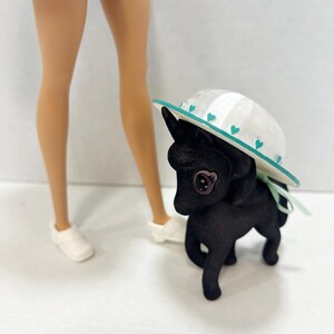 May include: A black toy pony with a textured surface and a large button eye, wearing a white sun hat with a teal brim and small teal heart accents. The pony is positioned next to the lower legs of a doll wearing white shoes and a patterned skirt.
