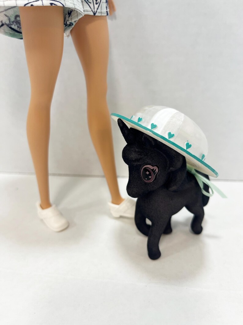 May include: A black toy pony with a textured surface and a large button eye, wearing a white sun hat with a teal brim and small teal heart accents. The pony is positioned next to the lower legs of a doll wearing white shoes and a patterned skirt.