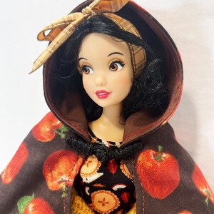 Op de afbeelding: Een pop met zwart haar draagt een bruine cape met capuchon met een rode appelprint en een beige geruite strik. De pop draagt een zwarte jurk met een patroon van appelschijfjes en een gele rok eronder. De cape heeft een zwarte sluiting.
