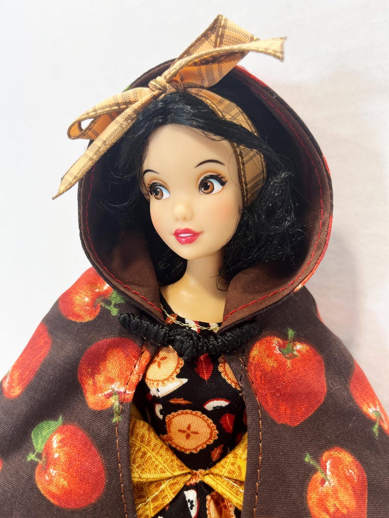 Op de afbeelding: Een pop met zwart haar draagt een bruine cape met capuchon met een rode appelprint en een beige geruite strik. De pop draagt een zwarte jurk met een patroon van appelschijfjes en een gele rok eronder. De cape heeft een zwarte sluiting.