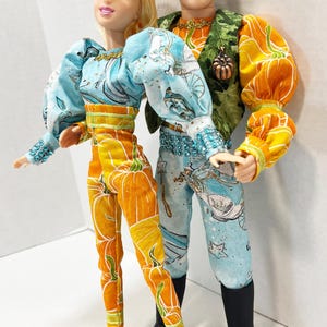 以下が含まれることがあります： ファンタジーコスチュームを着た2体の人形。1体は白い模様のライトブルーのトップスとオレンジ色のカボチャのズボンを着用。もう1体は緑色のベストとオレンジ色のカボチャの袖、ライトブルーのズボンを着用。どちらの人形もアクセサリーを身につけています。
