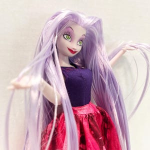 Pu&ograve; includere: Una bambola con lunghi capelli lisci color viola chiaro, occhi verdi e labbra rosse. La bambola indossa un top senza maniche viola scuro con sottili cuciture rosse sullo scollo e una gonna testurizzata rosa e rossa con motivo marmorizzato.