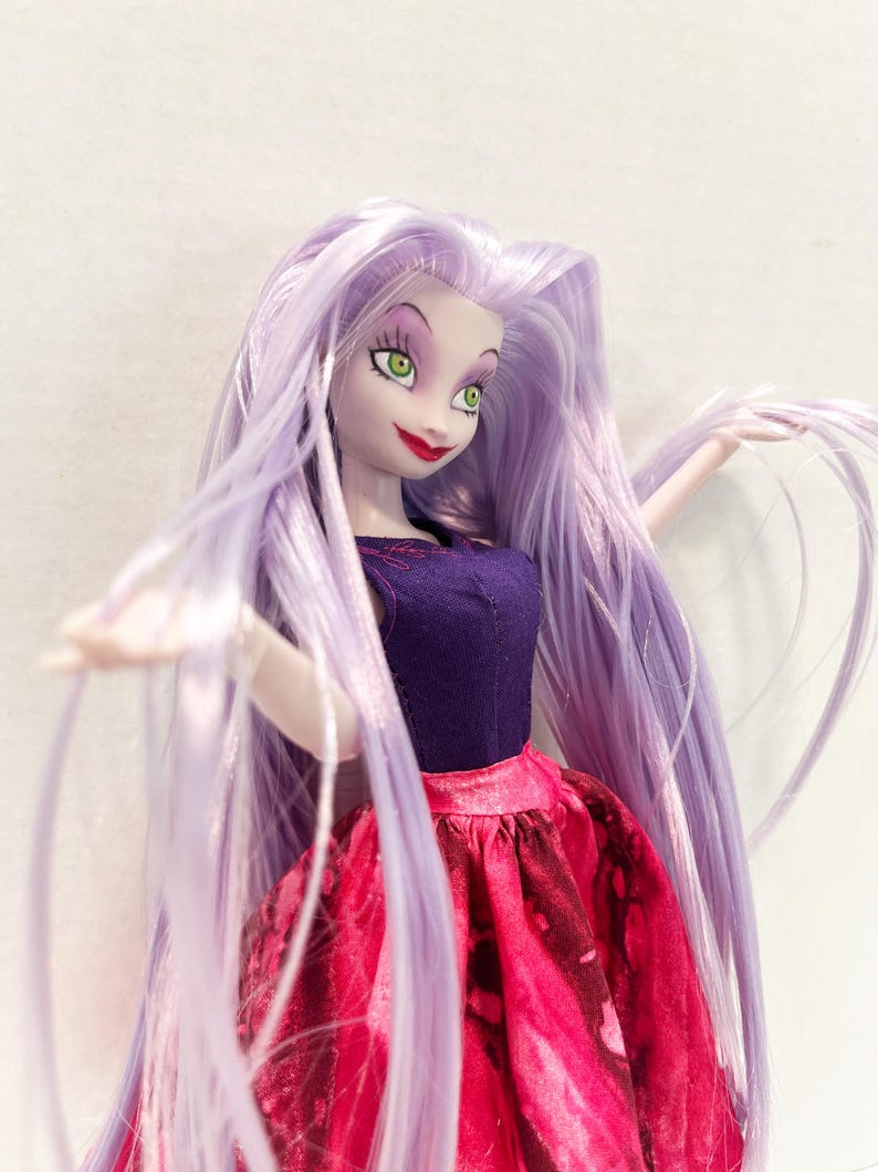Pu&ograve; includere: Una bambola con lunghi capelli lisci color viola chiaro, occhi verdi e labbra rosse. La bambola indossa un top senza maniche viola scuro con sottili cuciture rosse sullo scollo e una gonna testurizzata rosa e rossa con motivo marmorizzato.