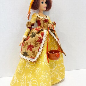 May include: A doll with auburn hair wearing a straw hat with a yellow flower and a long dress. The dress features a yellow skirt with white line drawings of pumpkins and leaves, and a brown bodice overlay with a red and brown maple leaf pattern.