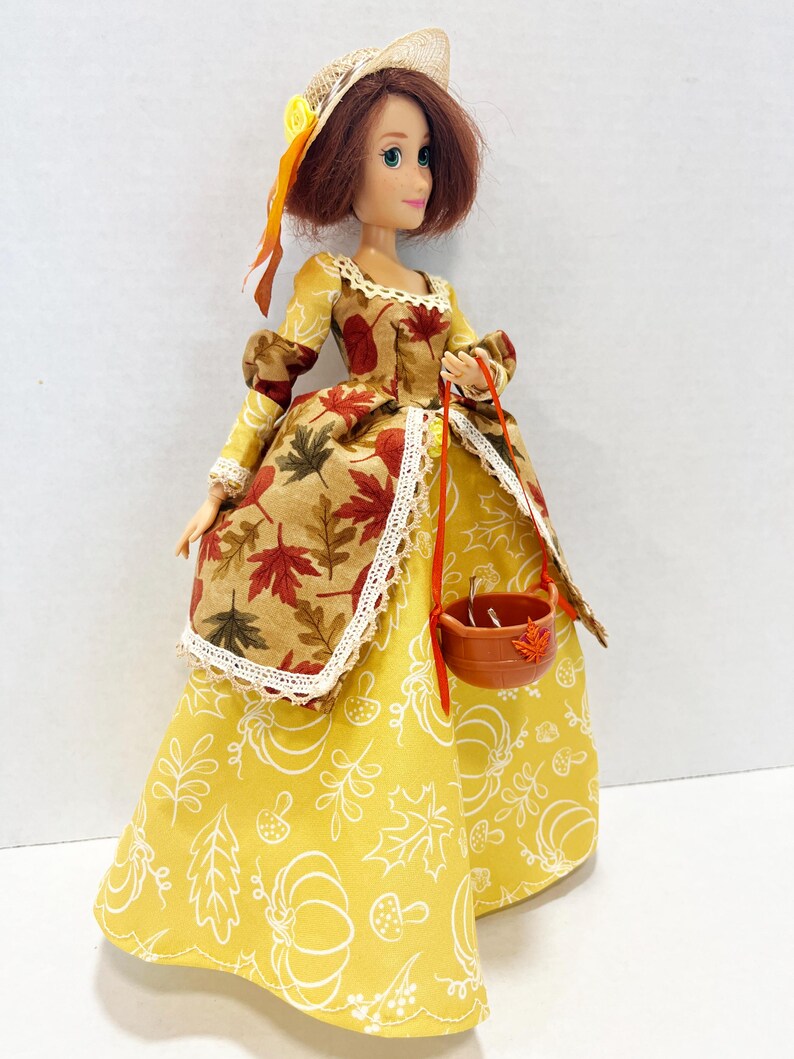May include: A doll with auburn hair wearing a straw hat with a yellow flower and a long dress. The dress features a yellow skirt with white line drawings of pumpkins and leaves, and a brown bodice overlay with a red and brown maple leaf pattern.