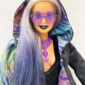 May include: A fashion doll with long, blue and silver hair, wearing purple-tinted sunglasses and a black hooded jacket with a colorful patterned lining. The doll also has a purple necklace and black lipstick.