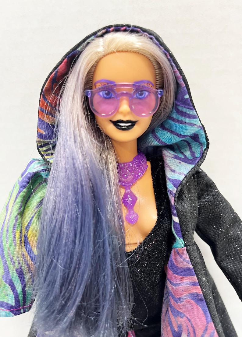May include: A fashion doll with long, blue and silver hair, wearing purple-tinted sunglasses and a black hooded jacket with a colorful patterned lining. The doll also has a purple necklace and black lipstick.