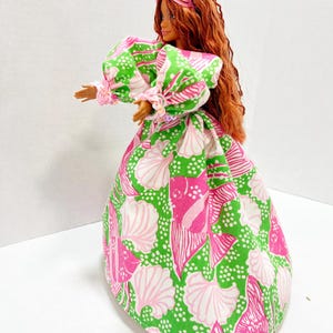 May include: A doll wearing a vibrant, patterned dress with a seashell design in pink and white on a green background. The dress has puffy sleeves and a full skirt. The doll has long, red hair and a pink headpiece.