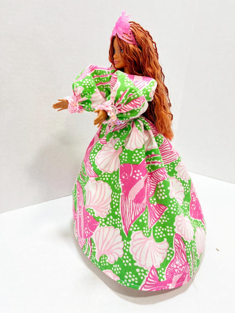 May include: A doll wearing a vibrant, patterned dress with a seashell design in pink and white on a green background. The dress has puffy sleeves and a full skirt. The doll has long, red hair and a pink headpiece.
