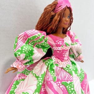 May include: A doll wearing a pink, green, and white patterned dress with puff sleeves and a matching pink headpiece. The doll has brown hair and a sequined bodice.