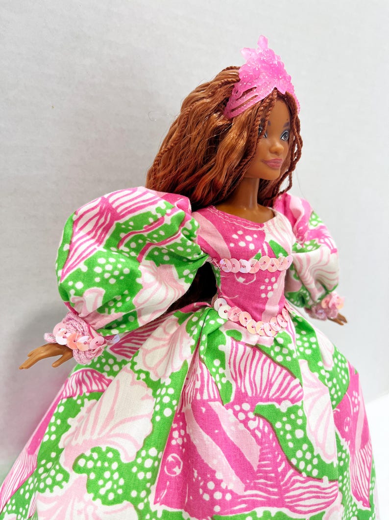 May include: A doll wearing a pink, green, and white patterned dress with puff sleeves and a matching pink headpiece. The doll has brown hair and a sequined bodice.