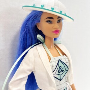 May include: A doll with long blue hair wearing a white hat with teal heart accents, a white cropped jacket, and a white dress featuring a grid pattern, teal geometric shapes, and a dark blue ampersand symbol on the bodice with light green straps.