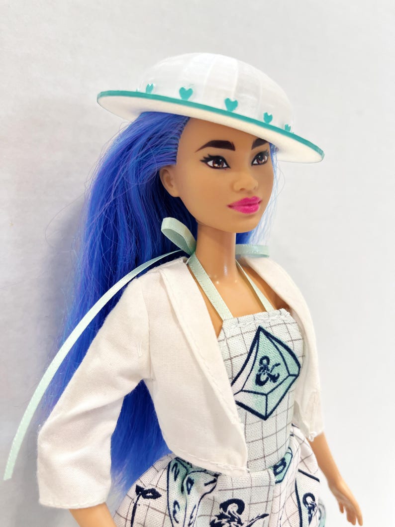 May include: A doll with long blue hair wearing a white hat with teal heart accents, a white cropped jacket, and a white dress featuring a grid pattern, teal geometric shapes, and a dark blue ampersand symbol on the bodice with light green straps.