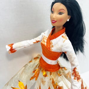 May include: A doll with long black hair and brown eyes wearing a white long-sleeved top with orange leaf patterns, an orange bodice with a white waistband, and a full skirt featuring a sheer overlay decorated with metallic orange autumn leaves.
