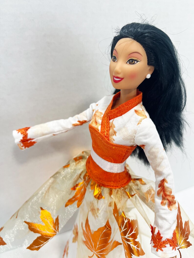 May include: A doll with long black hair and brown eyes wearing a white long-sleeved top with orange leaf patterns, an orange bodice with a white waistband, and a full skirt featuring a sheer overlay decorated with metallic orange autumn leaves.