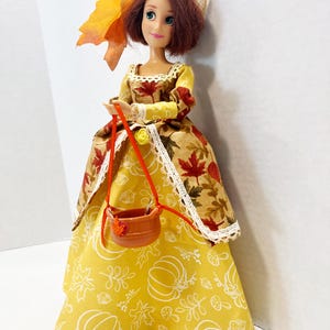 May include: A doll with auburn hair wearing a tan hat with an orange leaf accent. The doll wears a dress with a bodice featuring a brown autumn leaf print and a yellow skirt with white line drawings of pumpkins, leaves, and mushrooms. A small brown basket hangs from the doll's hands.