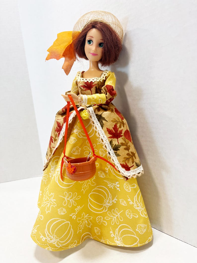 May include: A doll with auburn hair wearing a tan hat with an orange leaf accent. The doll wears a dress with a bodice featuring a brown autumn leaf print and a yellow skirt with white line drawings of pumpkins, leaves, and mushrooms. A small brown basket hangs from the doll's hands.