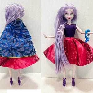 Pu&ograve; includere: Una bambola con lunghi capelli color lavanda indossa un top smanicato viola scuro, una gonna svasata rosa acceso e un mantello blu con un motivo floreale scuro. La bambola ha scarpe con tacco viola e tiene in mano un piccolo accessorio blu.