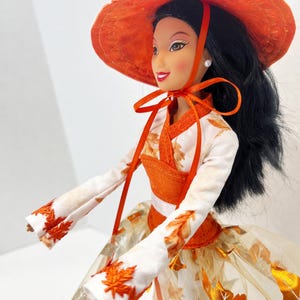 May include: A doll with long black hair wearing a wide-brimmed orange hat tied with a ribbon. The doll wears a white long-sleeved top with orange leaf embroidery and a matching skirt featuring a metallic orange leaf pattern and a sheer overlay.