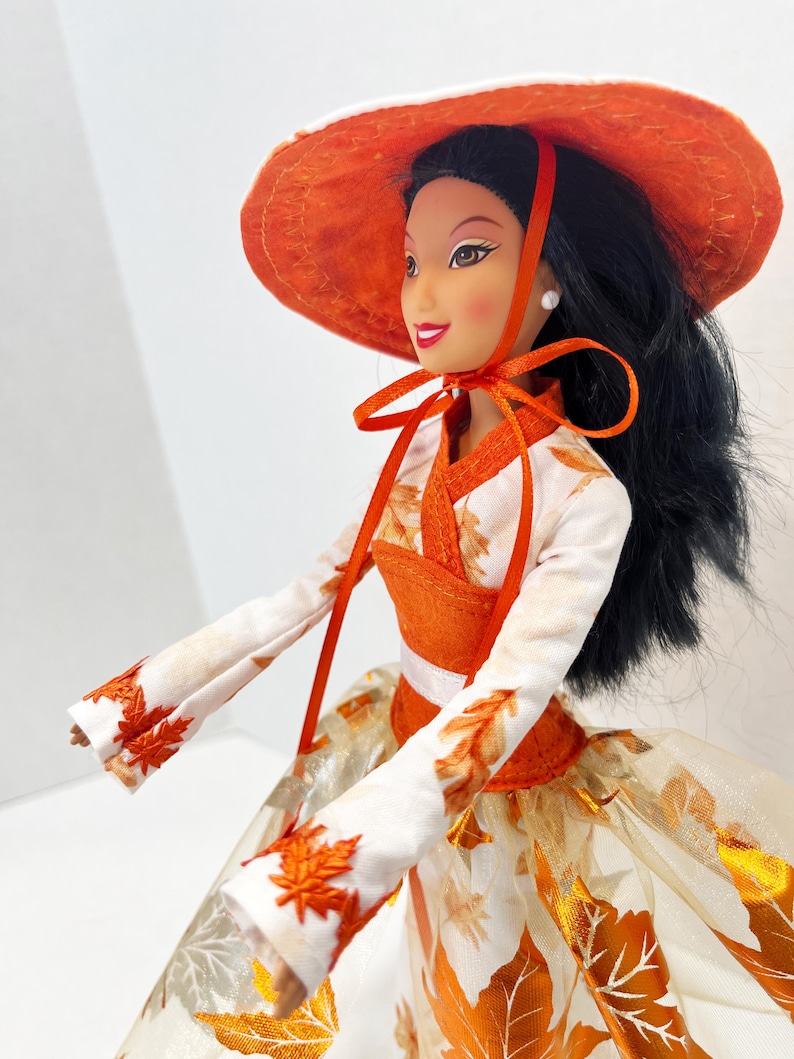 May include: A doll with long black hair wearing a wide-brimmed orange hat tied with a ribbon. The doll wears a white long-sleeved top with orange leaf embroidery and a matching skirt featuring a metallic orange leaf pattern and a sheer overlay.
