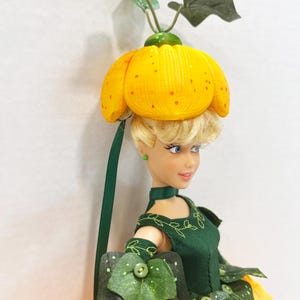 May include: A doll with blonde hair wearing a yellow pumpkin-shaped hat with green leaves on top. The doll wears a dark green dress with embroidered vine details, green leaf-shaped accents, and a yellow skirt. The doll has blue eyes and green earrings.