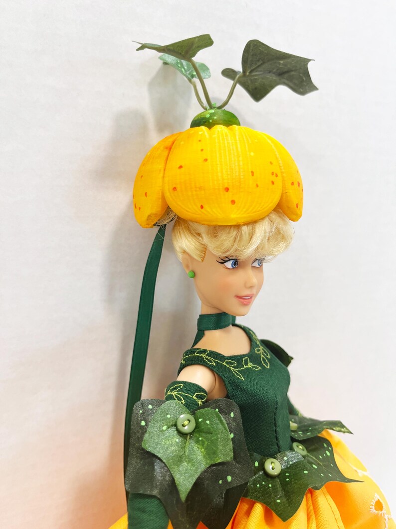 May include: A doll with blonde hair wearing a yellow pumpkin-shaped hat with green leaves on top. The doll wears a dark green dress with embroidered vine details, green leaf-shaped accents, and a yellow skirt. The doll has blue eyes and green earrings.