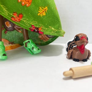 K&ouml;nnte beinhalten: Eine Puppe tr&auml;gt einen gr&uuml;nen Rock mit Herbstblattmuster und hellgr&uuml;ne Plateausandalen, begleitet von einer kleinen braunen Truthahnfigur mit rotem Kehllappen und einem h&ouml;lzernen Miniatur-Nudelholz mit einer d&uuml;nnen gr&uuml;nen Schnur.