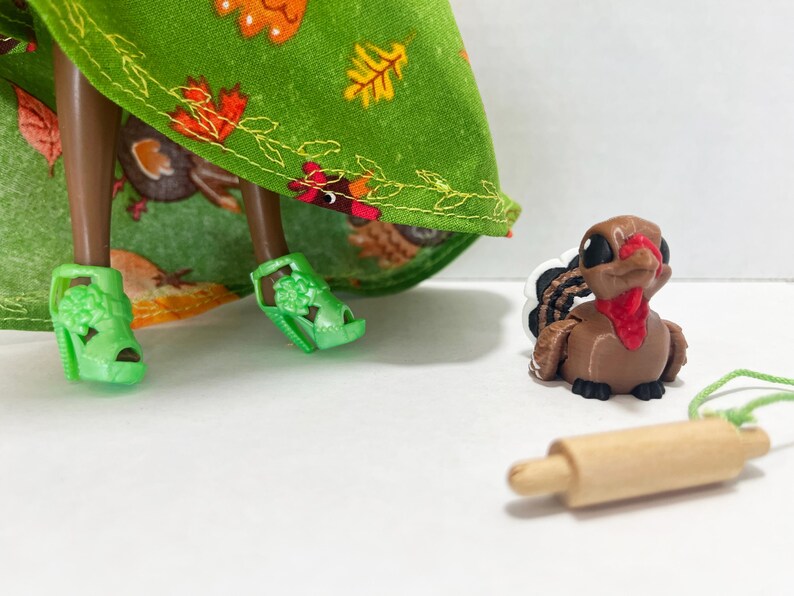 K&ouml;nnte beinhalten: Eine Puppe tr&auml;gt einen gr&uuml;nen Rock mit Herbstblattmuster und hellgr&uuml;ne Plateausandalen, begleitet von einer kleinen braunen Truthahnfigur mit rotem Kehllappen und einem h&ouml;lzernen Miniatur-Nudelholz mit einer d&uuml;nnen gr&uuml;nen Schnur.