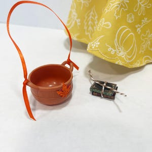 May include: A brown plastic bucket with a bright orange ribbon handle and a small orange leaf embellishment, accompanied by two miniature stacked books tied with a thin string, positioned near a yellow fabric with a white autumn-themed print.