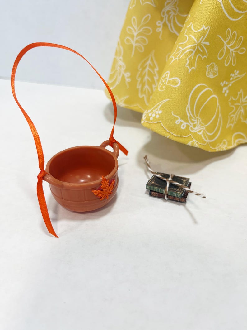 May include: A brown plastic bucket with a bright orange ribbon handle and a small orange leaf embellishment, accompanied by two miniature stacked books tied with a thin string, positioned near a yellow fabric with a white autumn-themed print.