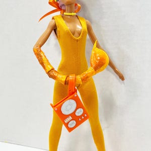 K&ouml;nnte beinhalten: Eine Puppe tr&auml;gt einen gelben &auml;rmellosen Jumpsuit, gelbe Kopfh&ouml;rer und eine violette Sonnenbrille. Die Puppe hat einen strukturierten orangefarbenen Armaufsatz, ein gelbes Halsband und h&auml;lt ein leuchtend orangefarbenes quadratisches Zubeh&ouml;rteil.
