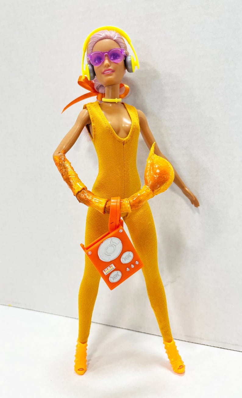 K&ouml;nnte beinhalten: Eine Puppe tr&auml;gt einen gelben &auml;rmellosen Jumpsuit, gelbe Kopfh&ouml;rer und eine violette Sonnenbrille. Die Puppe hat einen strukturierten orangefarbenen Armaufsatz, ein gelbes Halsband und h&auml;lt ein leuchtend orangefarbenes quadratisches Zubeh&ouml;rteil.