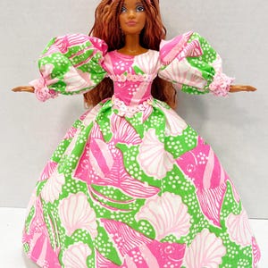 May include: A doll wearing a pink tiara and a long gown with a seashell and fish pattern in pink, white, and green. The doll has long brown hair and is wearing a pink and white dress with puffy sleeves.