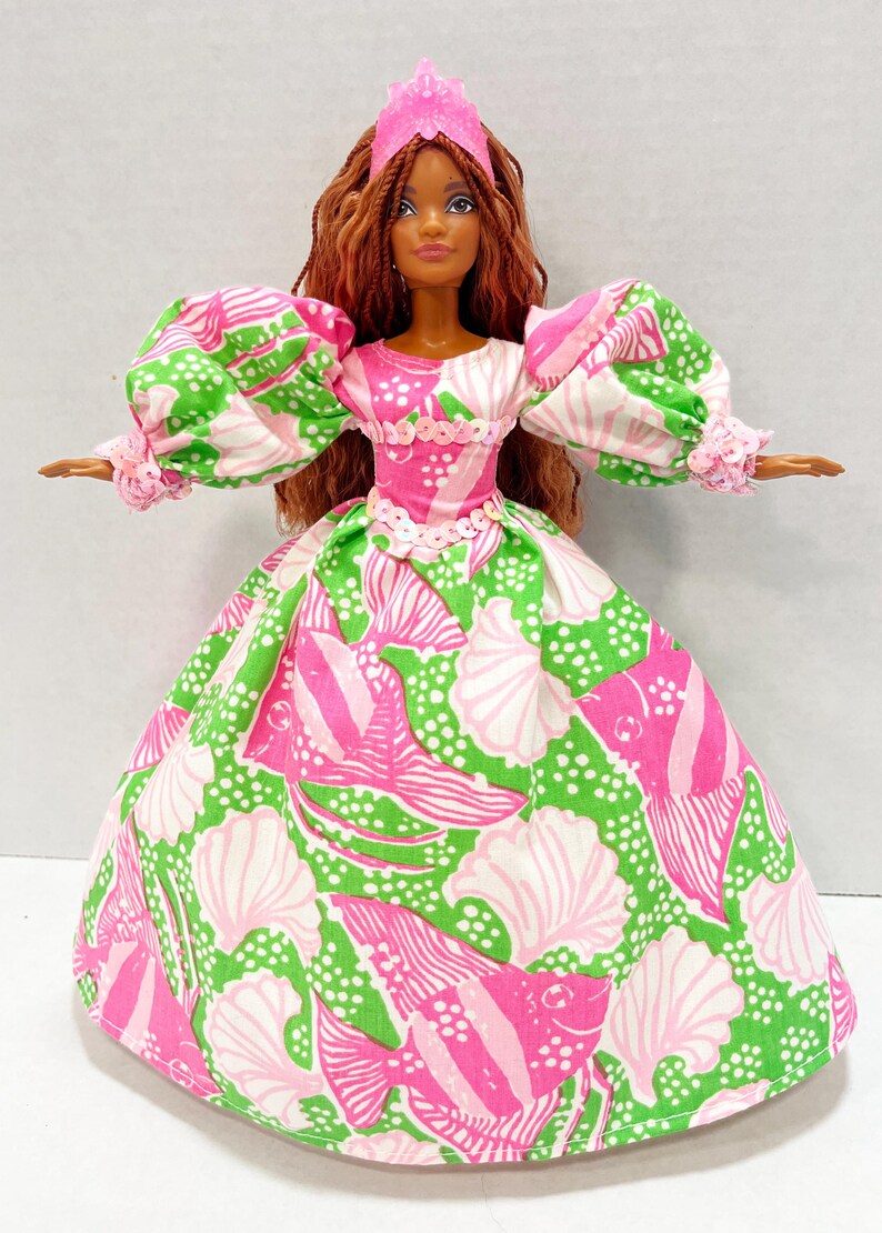 May include: A doll wearing a pink tiara and a long gown with a seashell and fish pattern in pink, white, and green. The doll has long brown hair and is wearing a pink and white dress with puffy sleeves.